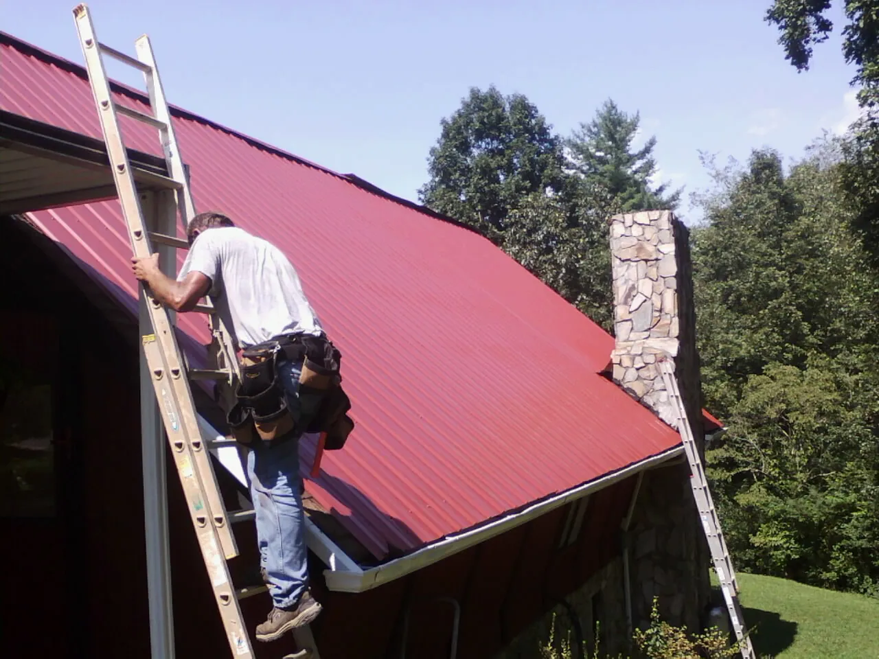 Roofer on a ladder beside a red metal roof and stone chimney — Soffit & Fascia Repair by Roofing Brothers in Guinda